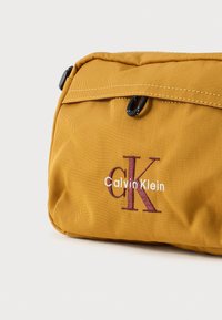 Žltá nylonová taška s prekrytím v hornej časti. Obsahuje vyšívané logo "Calvin Klein" v bielej a tmavočervenej farbe, s čiernym zipsom a doplnkami.