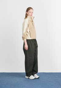 Top de fleece em bege e creme com zíper até à metade, com um bolso frontal, combinado com calças cargo verdes escuras e ténis brancos.