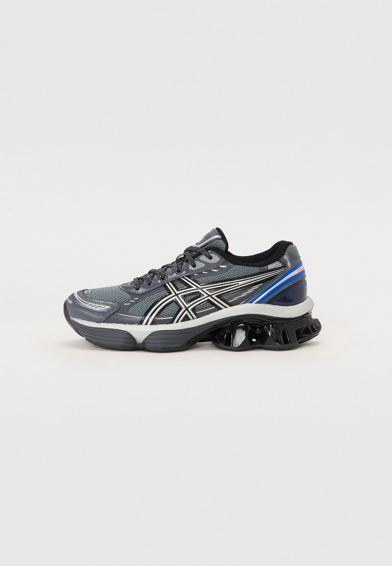 Zapatilla deportiva con parte superior de malla gris y acentos en negro y plata, una entresuela blanca gruesa y una suela exterior con patrón negro y azul.