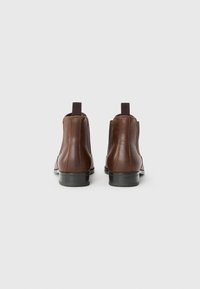 Botas de tobillo de cuero marrón con paneles laterales elásticos, tiradores y un tacón negro bajo. Textura suave, diseño minimalista.