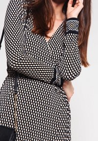 Black wrap dress with white and gold dot pattern, featuring long sleeves with black and white geometric cuffs, and a fitted silhouette.