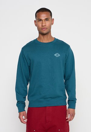 Mann trägt ein türkisfarbenes Sweatshirt mit Rundhalsausschnitt und kleinem weißem Logo auf der Brust sowie rote Hose, steht vor einem einfarbigen hellen Hintergrund.