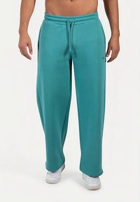 Grønne joggingbukser med snøre i taljen, tilspidsede ben og sidelommer, lavet af blødt stof. Hvide sneakers på fødderne.