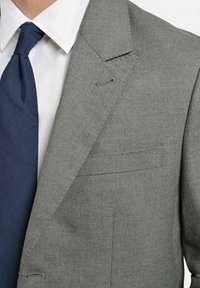 Grå ullblazer med öppna slag, en bröstficka och synlig sömnad. Kombinerad med en mörkblå slips och vit skjorta.