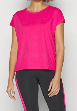 Kobieta w jasnoróżowym krótkim topie i czarnych legginsach z różowymi paskami po bokach, stoi z rękami swobodnie opartymi wzdłuż ciała.