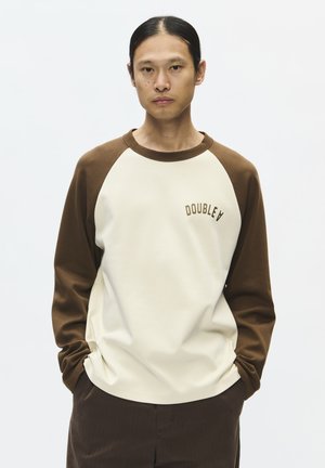 Ung person med langt sort hår samlet bagud iført creme og brun sweatshirt med teksten "DOUBLE A" og mørkebrune bukser, hænderne i lommerne.