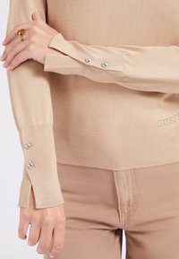 Beżowa bluzka z długim rękawem z srebrnymi guzikami na mankietach, noszona z pasującymi beżowymi spodniami oraz dłonią prezentującą bursztynowy pierścionek.