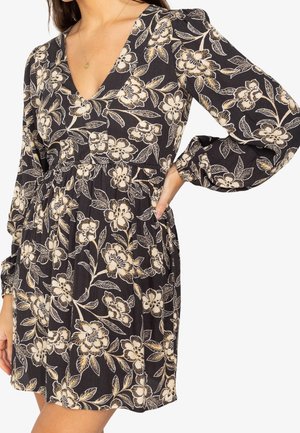 Czarna sukienka z beżowym kwiatowym nadrukiem, długie bufiaste rękawy, dekolt w kształcie litery V i marszczona talia, noszona przez osobę z ręką na biodrze.
