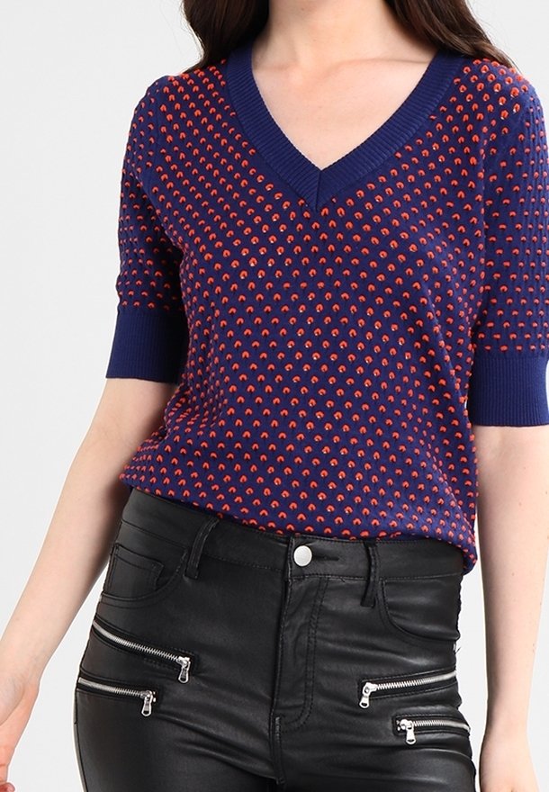 Námořnický svetr s krátkým rukávem a výstřihem do V, zdobený červeným obloukovým vzorem a žebrovanými manžetami. Nosený s černými kalhotami z imitace kůže se zipy.