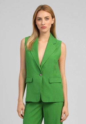 Femme portant un blazer vert sans manches avec un seul bouton et un pantalon vert assorti, debout devant un fond gris uni.