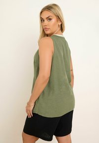 Vihreäoliivinvärinen, hihaton toppi pyöreällä kaula-aukolla, jossa on valkoinen aaltoileva yksityiskohta takana, yhdistetty mustiin shortseihin.