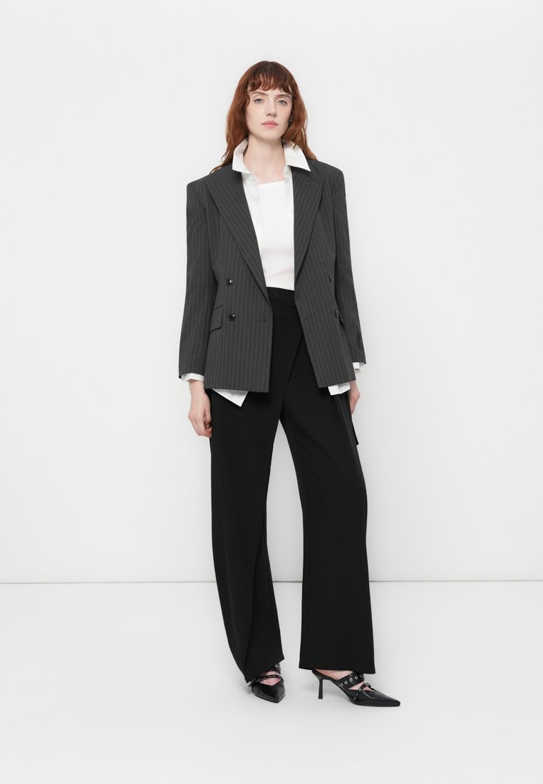 Blazer gris à double boutonnage et rayures verticales, porté sur une chemise blanche, associé à un pantalon noir à jambes larges et des chaussures noires à talons avec des découpes.