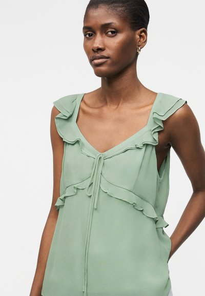 Νεαρή γυναίκα με κοντά μαλλιά που φορά ανοιχτοπράσινο αμάνικο μπλουζάκι με βολάν και ζώνη, στέκεται μπροστά σε απλό φόντο.