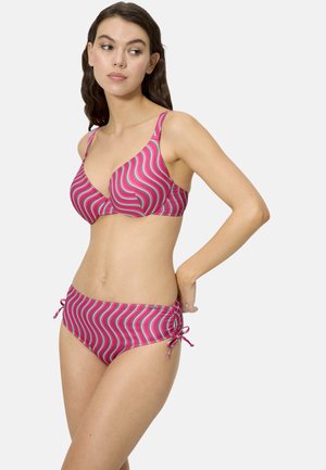 Mujer con cabello largo y oscuro, vestida con un bikini de rayas onduladas en rosa y verde, ajustándose el lateral de la parte inferior sobre un fondo neutro.