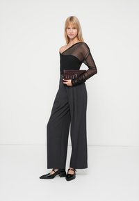 Top in rete nero con una spalla, abbinato a pantaloni grigi a gamba larga a vita alta. Tiene in mano una borsa a tracolla bordeaux scuro con dettagli metallici.