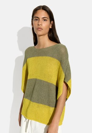 Młoda kobieta z długimi, ciemnymi włosami, nosząca luźny sweter z szerokimi, poziomymi paskami w kolorze oliwkowej zieleni i żółtym, stojąca na białym tle.