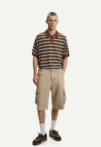 Polo a righe marroni con righe blu, pantaloni cargo beige, calze bianche e scarpe marroni. Vestibilità comoda, maniche corte e dettagli con bottoni.