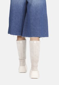 Knee-high batai iš smėlio spalvos zomšo, su apvaliu logotipo akcentu, derinami su nusidėvėjusiu džinsiniu sijonu, pabrėžiančiu laisvą siluetą.
