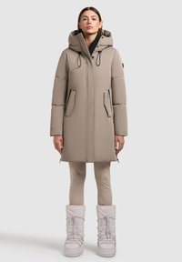 Beige puffer coat with adjustable hood, two front pockets, and elastic drawstrings. Paired with beige leggings and light-colored winter boots.
