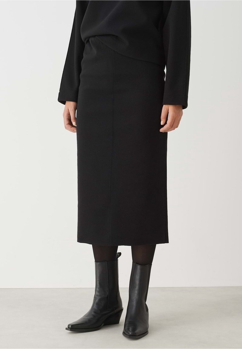Femme portant une jupe crayon longue noire, un haut à manches longues noir, des collants noirs et des bottines en cuir noir, debout sur un sol gris.