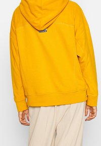 Gul huvtröja i mjukt material, med ribbad nederkant och ärmslut, samt en liten svart logotypetikett på baksidan.