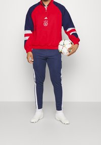 Chaqueta atlética roja y azul marino con medio zipper y logotipo, combinada con pantalones azul marino que tienen rayas blancas a los lados. Sosteniendo un balón de fútbol multicolor.