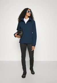 Tamsiai mėlyna vandeniui atspari striukė su aukštu apykakliu, priderintos aptemptos juodos kelnės ir sportbačiai, laiko juodą šalmą, modelis dėvi akinius nuo saulės.