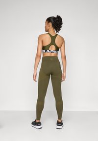 Olíva zöld sportmelltartó és leggings, racerback kivitelben; textúrált anyag; logó a derékszíjon; fekete sportcipővel párosítva.