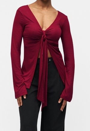 Femme portant un haut rouge profond à manches longues avec un nœud à l'avant et un col en V, associé à un pantalon noir sur un fond uni.