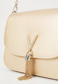 Sac à main texturé doré avec un rabat avant, présentant un fermoir métallique en forme de 'V' et un accent de gland en chaîne. Fini lisse et quincaillerie polie.