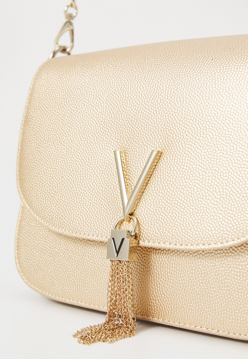 Sac à main texturé doré avec un rabat avant, présentant un fermoir métallique en forme de 'V' et un accent de gland en chaîne. Fini lisse et quincaillerie polie.