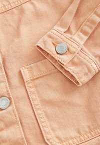 Lähikuvassa näkyvä persikkavärinen denimpaita, jossa on teksturoitu kangas, hopeanväriset nappikoristeet ja siististi ommeltu tasku.