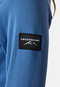 Blå langærmet skjorte med en tekstureret overflade og et sort patch, der viser "SWEDEMOUNT" og bjerggrafik på venstre ærme.