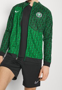 Chaqueta verde con patrón y cremallera, con acentos negros y un logo redondo, combinada con pantalones cortos negros que presentan el logo de Nike en blanco. Material texturizado.