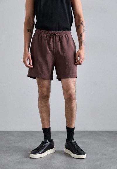 Braune Shorts mit elastischem Bund und Kordelzug, getragen mit einem schwarzen ärmellosen Oberteil und schwarzen Turnschuhen mit einer weißen Sohle.