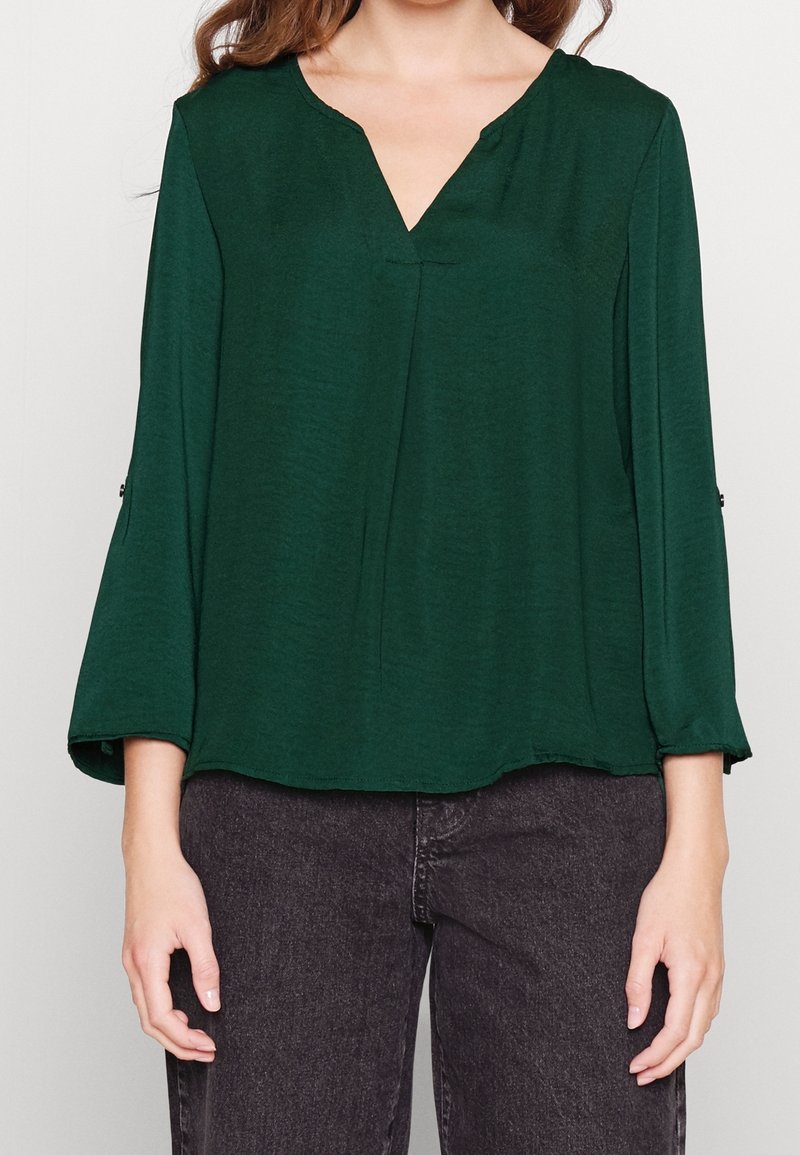 Kvinna iklädd en mörkgrön V-ringad blus med 3/4 ärmar och svarta jeans, stående med avslappnade armar vid sidorna mot en enfärgad bakgrund.