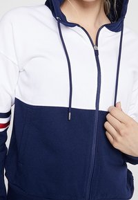 Sweat à capuche zippé marine et blanc avec un col haut, un cordon de serrage, des poches latérales et des accents de rayures rouge, blanc et bleu sur les manches. Texture de tissu douce.