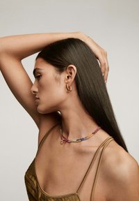 Pendientes de aro de oro, collar de cuentas coloridas con un charm, y una blusa marrón texturizada. Cabello oscuro lacio peinado hacia un lado.