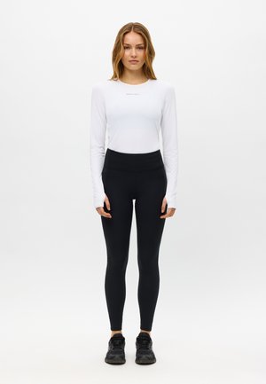 Junge Frau steht mit dem Gesicht nach vorne, trägt ein weißes Langarmshirt, schwarze hoch taillierte Leggings und schwarze Sneakers vor weißem Hintergrund.