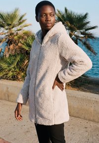 Moteris, vilkinti smėlio spalvos vilnonę striukę su užtrauktuku, stovi prie palmės medžių ir vandenyno, žvelgdama į šalį, viena ranka ant klubų.