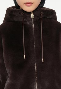 Haut à capuche en fausse fourrure marron avec un zip à l'avant, une capuche à cordon de serrage et des accents métalliques dorés. Texture douce et apparence moelleuse.