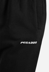 Pantaloni sportivi neri realizzati in tessuto morbido, con una tasca laterale e il logo "PEGADOR" ricamato sulla coscia in un carattere audace.
