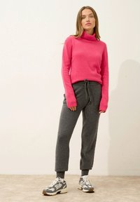 En varmt rosa polotröja med ribbade ärmslut, ihop med grå joggers med dragsko. Skorna är grå och vita sneakers med bruna detaljer.