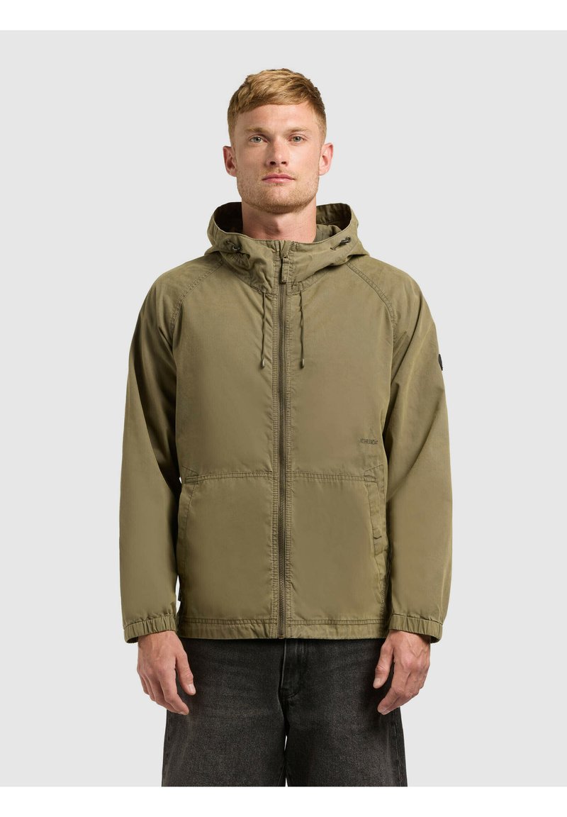 Mand iført olivengrøn hættetrøje med lynlås foran og sorte bukser, stående mod en ensartet lys baggrund.