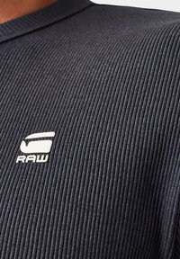 Svart ribbad t-shirt i tyg med rund halsringning och en liten vit "RAW"-logotyp på vänster sida. Texturerad yta med subtil lyster.