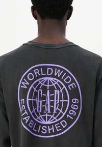 Juodas džemperis su dideliu levandų spalvos grafiniu dizainu, vaizduojančiu žemėlapį, ir užrašu "WORLDWIDE GAP ESTABLISHED 1969". Unisex dizainas.