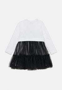 Bela obleka z dolgimi rokavi in črnim tilom na krilu, ki ima plastne naborke na robu za dodatno teksturo in volumen.