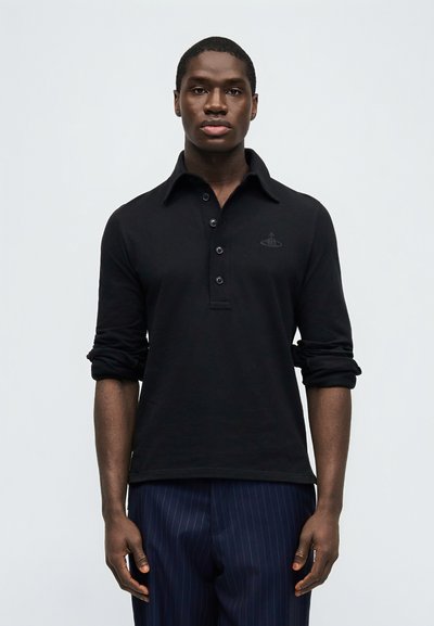 Άνδρας που φοράει μαύρο πόλο μπλουζάκι με μακριά μανίκια, κουμπιά και μανίκια γυρισμένα, σε συνδυασμό με μπλε σκούρο παντελόνι με ρίγες.