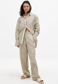Striped pajamas in beige and brown hues, featuring a button-up shirt with a relaxed fit and elastic waistband pants. Styled with slip-on sandals.