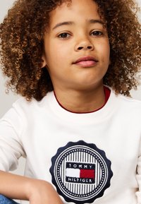 Felpa bianca con logo tondo blu navy che presenta il testo "TOMMY HILFIGER" e dettagli in rosso, bianco e blu; morbido tessuto di cotone. Vestibilità casual.